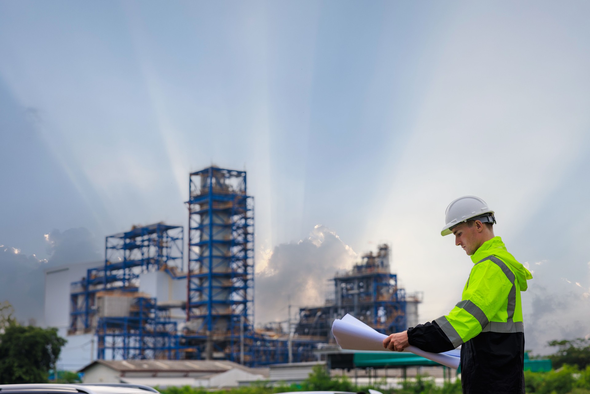 engineer in uniform and hardhat stood looking down at blueprint at oil refinery plant construction site and sunlight clouds sky backgrounds.